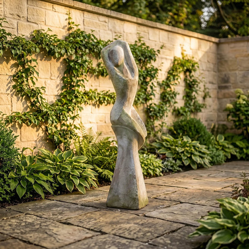 Statue of a abstract woman statue in a garden setting with a stone wall and plants.