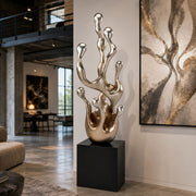 A metallic silver abstract sculpture with a black pedestal base, displayed in a modern loft-style living room next to a large abstract wall art piece.