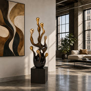 A modern abstract sculpture with metallic gold and dark bronze dripping forms on a black pedestal, displayed in a stylish loft apartment next to a large abstract painting.