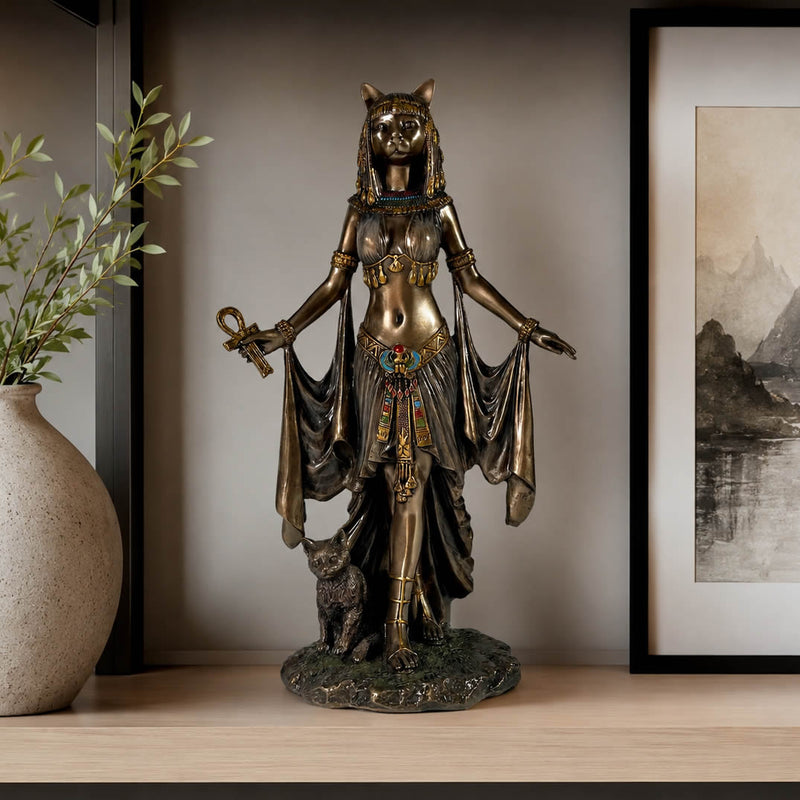 An Egyptian goddess statue with a cat head, holding an ankh and wearing a detailed gold and turquoise dress, standing on a base with a small cat, displayed on a wooden shelf next to a vase and framed picture. Bastet Egyptian Goddess Of Protection Statue