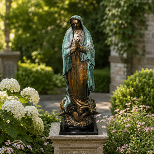 A bronze and green patina statue of the Virgin Mary standing on a stone pedestal in a garden setting with green foliage and white flowers.