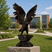 A dark bronze eagle sculpture with wings spread wide, perched on a textured rock base atop a stone pedestal, set in a landscaped lawn with a modern building in the background.