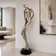 A tall, slender metallic silver sculpture of a stylized female figure standing on a black circular base, displayed in a modern living room next to a large abstract painting.