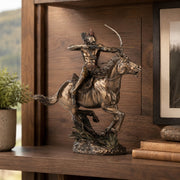 Bronze-finished sculpture of a Native American rider on a galloping horse, displayed on a wooden shelf next to a small potted plant and books.