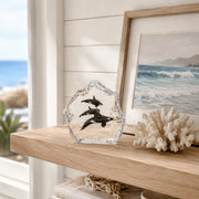 Decorative Lucite sculpture of Orca Whales on a wooden shelf with a framed beach scene painting in the background.