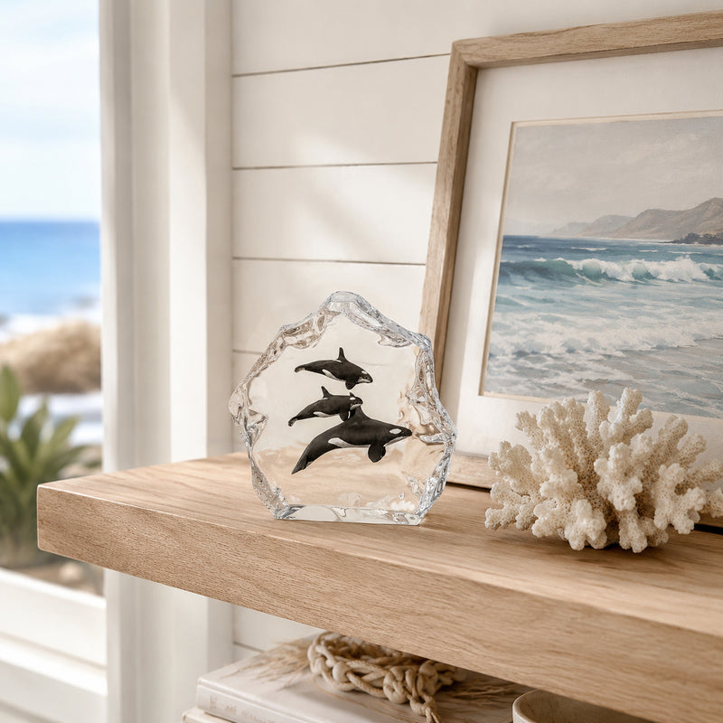 Decorative Lucite sculpture of Orca Whales on a wooden shelf with a framed beach scene painting in the background.