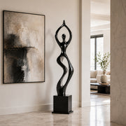 A glossy black and silver abstract sculpture of a stylized female figure standing on a rectangular pedestal, displayed in a modern interior hallway next to a large abstract wall painting.