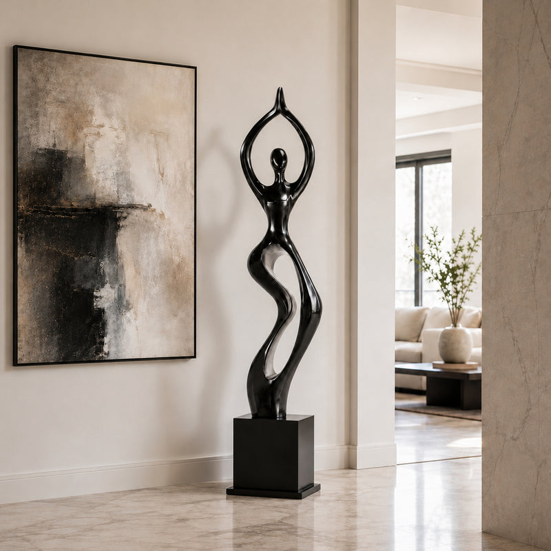 A glossy black and silver abstract sculpture of a stylized female figure standing on a rectangular pedestal, displayed in a modern interior hallway next to a large abstract wall painting.