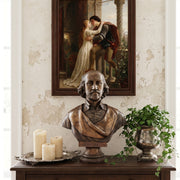 Bronze bust of William Shakespeare on a wooden table with candles and a painting of Romeo and Juliet in the background. 