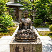 Bronze Buddha statue in a garden setting with greenery and traditional architecture.