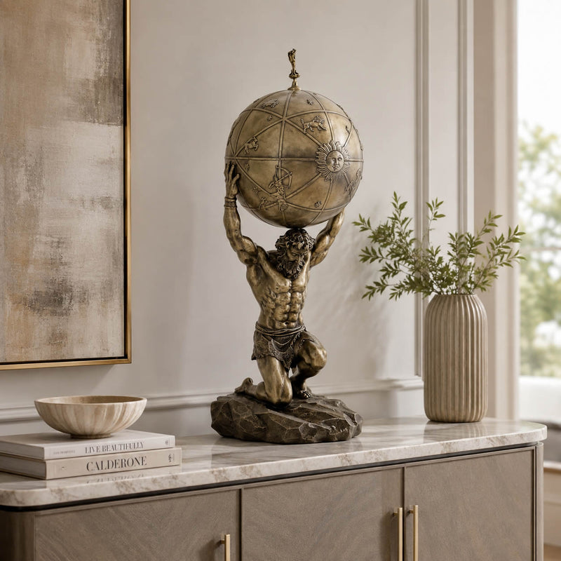 A bronze-finished sculpture of a muscular Atlas figure kneeling and supporting a detailed globe on his shoulders, displayed on a marble-topped console cabinet in a styled interior setting.