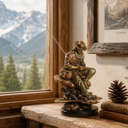 A bronze-finished sculpture of a seated fly fisherman on a log base, displayed on a rustic wooden shelf next to a window showing a mountain and pine forest view.