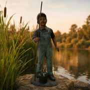 Bronze statue of a boy in overalls holding a fishing rod by a lake
