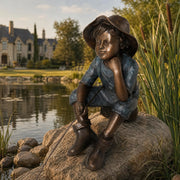 Bronze statue of a child sitting on a rock by a pond with a house in the background