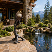 A large bronze eagle statue perched on a tree branch stands beside a stone patio and outdoor koi pond in a luxurious mountain chalet garden.