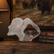 Decorative lucite sculpture of a buffalo bust inside of the sculpture are running buffalo. Placed a rustic wooden surface with books and a painting in the background.