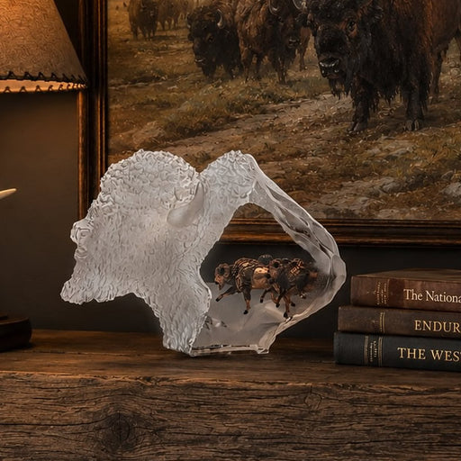 Decorative lucite sculpture of a buffalo bust inside of the sculpture are running buffalo. Placed a rustic wooden surface with books and a painting in the background.