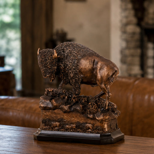 Bronze-finished bison figurine standing on a rocky base with a dark rectangular pedestal, displayed on a wooden table in a warm indoor setting.