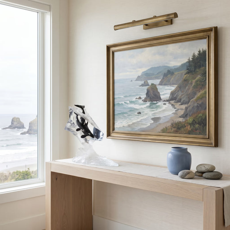 Framed artwork of a coastal scene on a wall above a wooden console table. Table has a orca whale sculpture on it