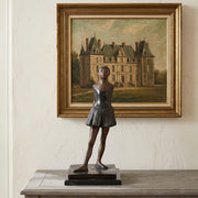 Bronze reproduction sculpture of Degas's Little Dancer of Fourteen Years on a table with a framed painting of a castle in the background.