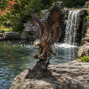 A detailed bronze eagle statue with wings spread, catching fish, set outdoors on a rock in front of a waterfall and pond.