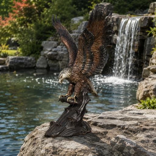 A detailed bronze eagle statue with wings spread, catching fish, set outdoors on a rock in front of a waterfall and pond.