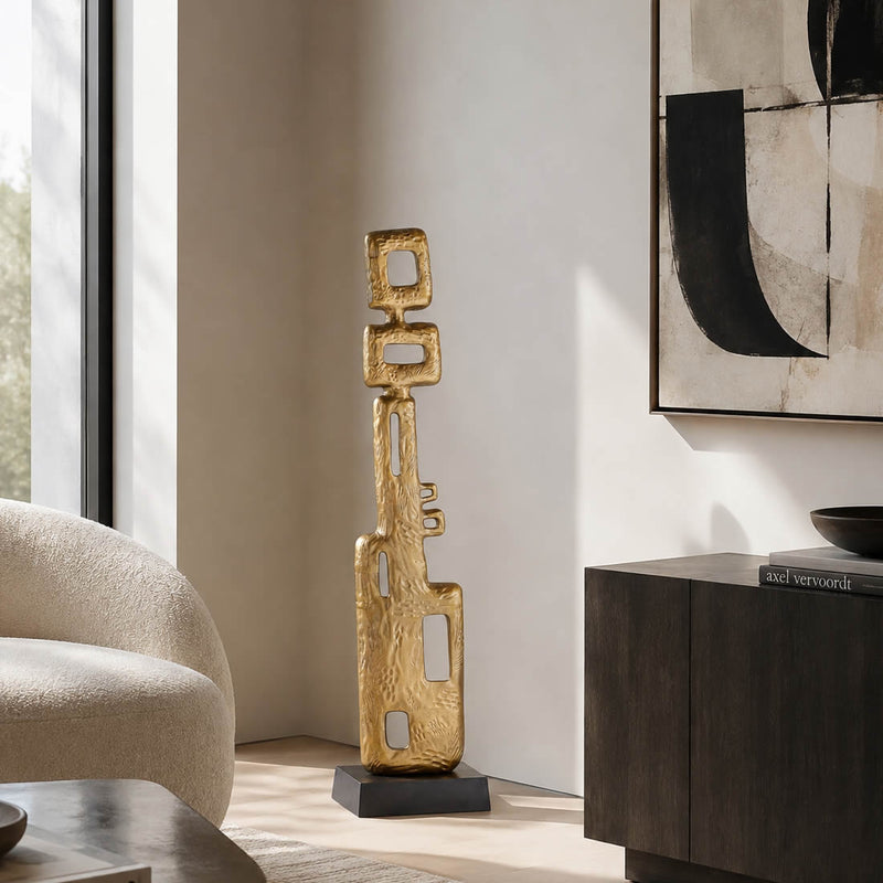 A tall, abstract gold metal sculpture with a textured finish stands on a black base in a modern living room corner next to a beige armchair and a dark wood sideboard.