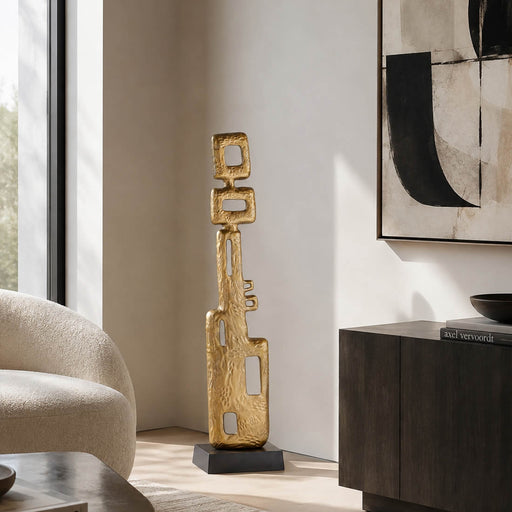 A tall, abstract gold metal sculpture with a textured finish stands on a black base in a modern living room corner next to a beige armchair and a dark wood sideboard.