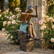 Bronze statue of a girl reading, sitting on a stack of books in a garden.