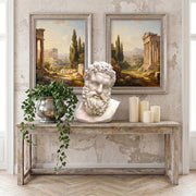 Decorative console table with classical bust of Hercules, candles, and potted plant against a wall with framed landscape paintings.