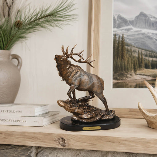 Bronze-colored elk statue standing on a rocky base with a black round pedestal, displayed on a wooden shelf in a styled home setting with books, a vase, and a framed mountain landscape print.