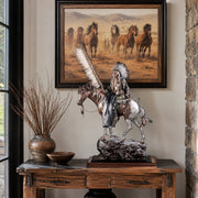 Decorative statue of a Native American on horseback on a wooden table with a framed painting of horses in the background.