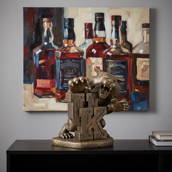 Decorative University of Kentucky Wildcats statue on a table in front of a painting o whiskey  bottles.