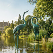 Two green metal heron statues by a pond with water flowing from their beaks.