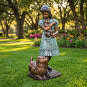 Bronze statue of a girl holding a cat and a dog on a grassy lawn with flowers and trees in the background