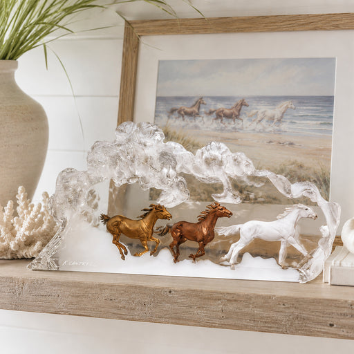 Decorative sculpture of horses running in lucite on a shelf with a framed beach scene in the background. Sculpture artist is Kitty Cantrell. Title of sculpture: Praire Storm