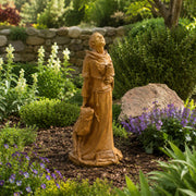 A garden statue of Saint Francis, robed  holding a book and a small animal, placed in a landscaped garden bed with mulch, shrubs, flowers, and a stone wall in the background.