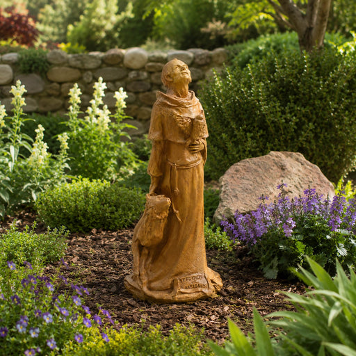A garden statue of Saint Francis, robed  holding a book and a small animal, placed in a landscaped garden bed with mulch, shrubs, flowers, and a stone wall in the background.