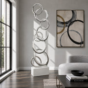 A tall, modern abstract sculpture made of interlocking metallic rings on a white rectangular base, displayed in a bright living room next to a window, with a gray sofa, framed wall art, and a coffee table nearby.
