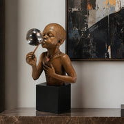 Bronze sculpture of a African American child blowing a bubble on a black cube pedestal against a neutral wall with abstract art. Title of sculpture is Summer Days. Artist: Thomas Blackshear