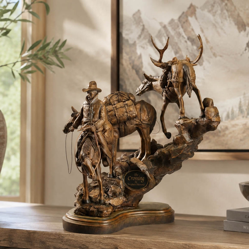 Bronze-finished decorative sculpture of a cowboy riding a pack mule with another deer on a rocky base, displayed on a wooden shelf in front of a framed mountain print.