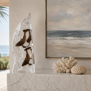 Decorative sculpture of humpback whales in clear acrylic on a marble surface with a coastal painting in the background.