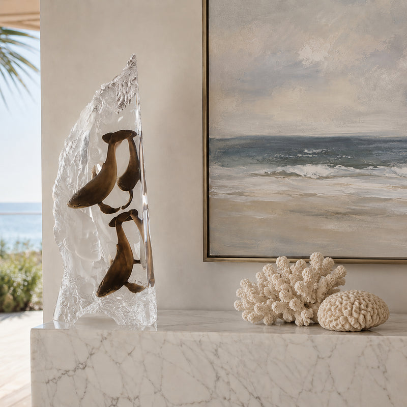 Decorative sculpture of humpback whales in clear acrylic on a marble surface with a coastal painting in the background.
