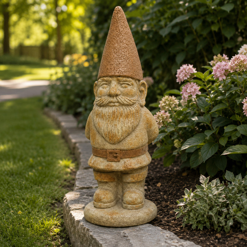 A stone garden gnome statue with a textured tan hat and beard, standing on a stone border in a garden with green foliage and pink flowers.